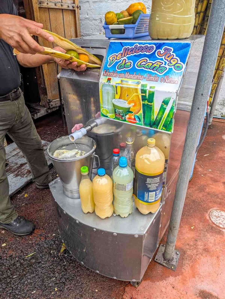 A street vendor puts two long pieces of sugar cane with whole oranges and limes in between into a machine to make sugarcane juice, a popular drink in Ecuador. A sign on the machine reads “Delicioso Jugo de Caña.”