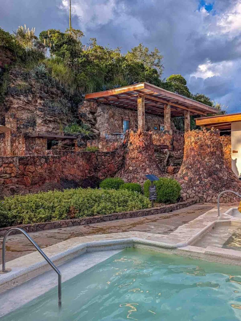 Outdoor pools at Piedra de Agua in Baños de Cuenca, set against a rocky hillside with lush greenery and dramatic clouds in the background.