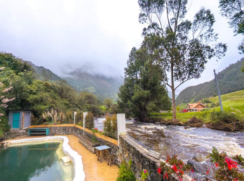 Scenic view of the thermal baths at Termas Pumamaqui in Cuenca Ecuador, with a stone-edged hot spring pool beside a rushing river and lush Andean mountains in the background.