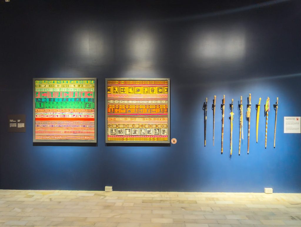 Colorful indigenous textiles and ceremonial staffs mounted on a dark blue wall inside the Pumapungo Museum in Cuenca.