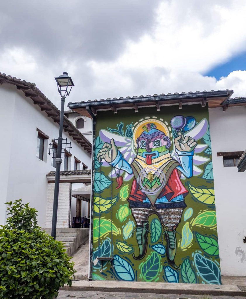 Vibrant street art mural inside Ciudad Mitad del Mundo, showcasing a figure wearing a Inti Raymi mask and elf like shoes with an ear of corn in the center of its chest.