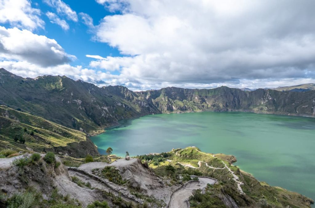 A panoramic view of Quilotoa Crater Lake filled with turquoise water under a partly cloudy sky, winding hiking trails visible in the foreground. This is one of the most iconic views on the Quilotoa Loop.