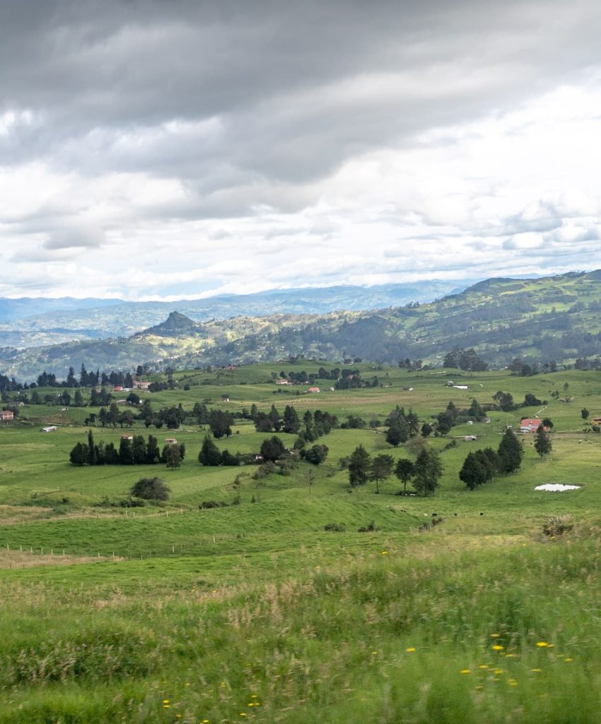 Scenic view of the Ecuadorian countryside with rolling green hills, scattered farmhouses, and dramatic clouds, as seen during a bus ride from Baños to Cuenca.