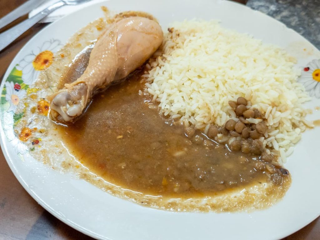 A plate of local Ecuadorian food with rice, lentils, and chicken drumstick in brown sauce, a common meal served at roadside stops on the bus from Baños to Cuenca.