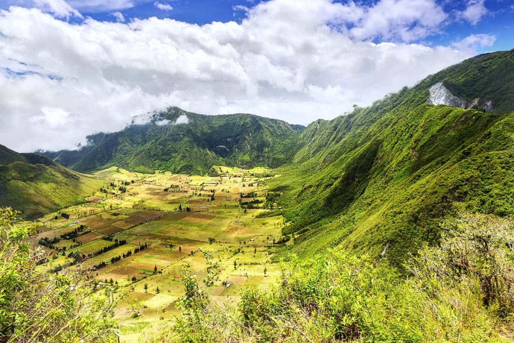 An ariel view of Pululahua Crater near Quito shows a patchwork of farm land with trees surrounded by lush green mountains under a blue sky with puffy clouds.