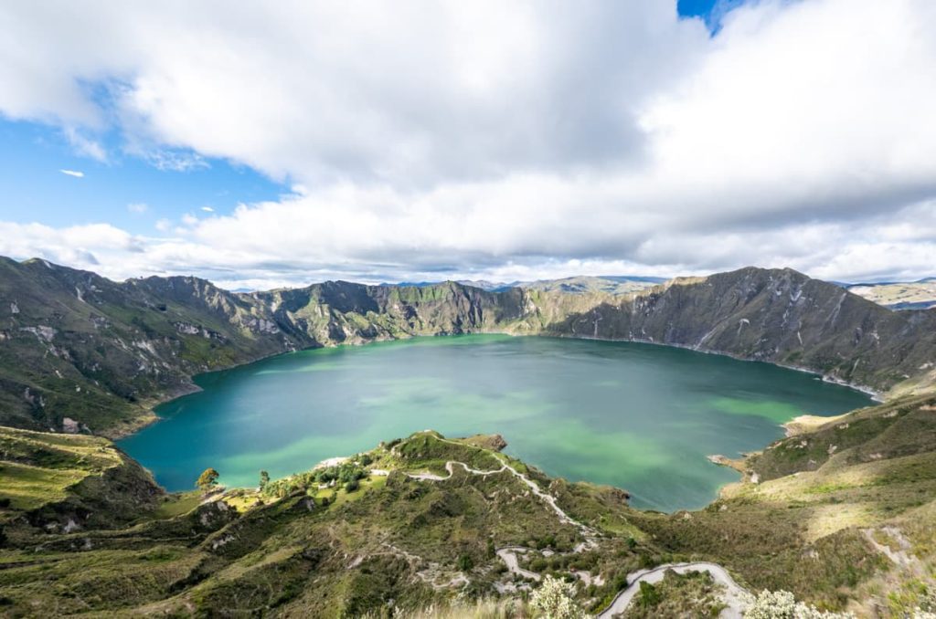 A wide panoramic view of the emerald-colored Quilotoa Lake from above, with winding paths leading to the crater lake shore.