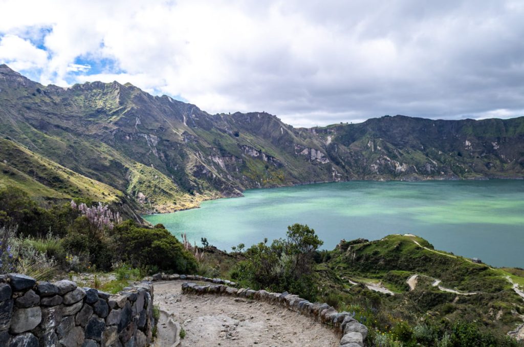A curving dirt trail bordered by stone walls winds down toward the calm, green waters of Laguna de Quilotoa, surrounded by lush mountains.