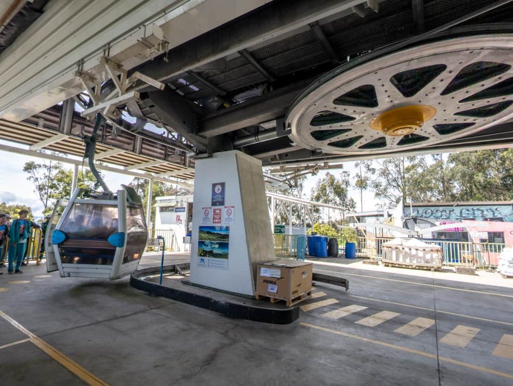 A cable car arrives at the boarding platform with visible machinery and signage at the lower station of the Teleférico de Quito.