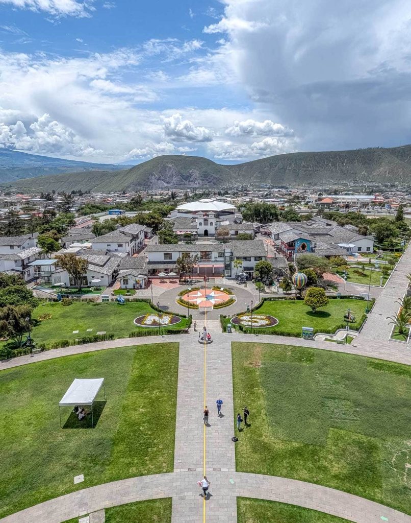 Aerial view of the Mitad del Mundo monument grounds with the yellow Ecuador equator line running through the city with buildings and mountains in the background.