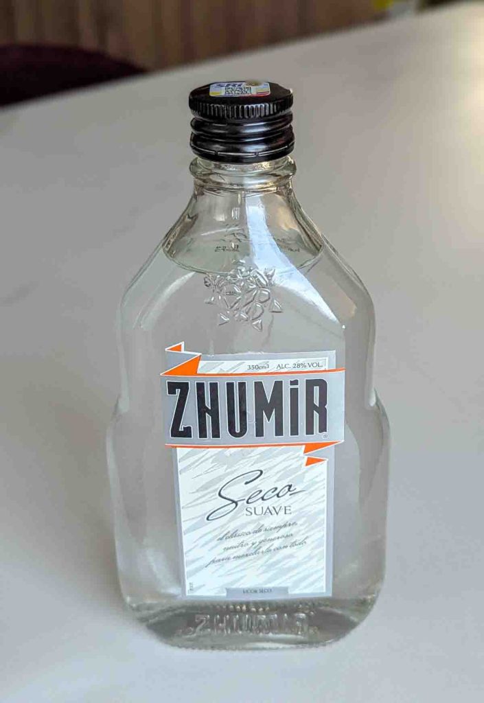 A clear glass bottle of Zhumir Seco Suave, a popular Ecuadorian alcoholic drink with 28% alcohol content, displayed on a white counter. The label features bold branding and text in Spanish.