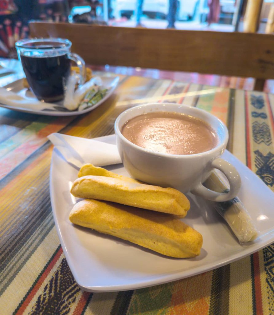 A white plate with three golden bizcochos, which look like short breadsticks. This traditional Ecuadorian snack is shown with a cup of hot chocolate and a cheese stick. The plate is sitting on a colorful Andean tablecloth. In the background another plate is seen with a cup of black coffee.