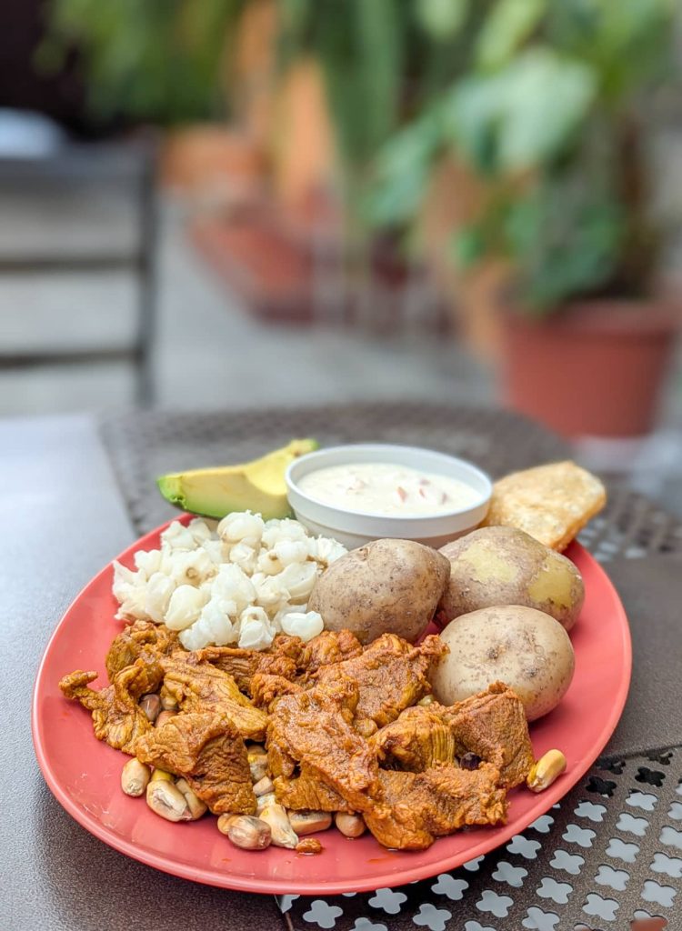 Pieces of carne colorada on top of crispy corn kernels, whole potatoes, mote, avocado, an empanada, and cheese sauce are served on a red oval plate. This vibrant dish from Cotacachi showcases the seasoning in traditional Ecuador food.