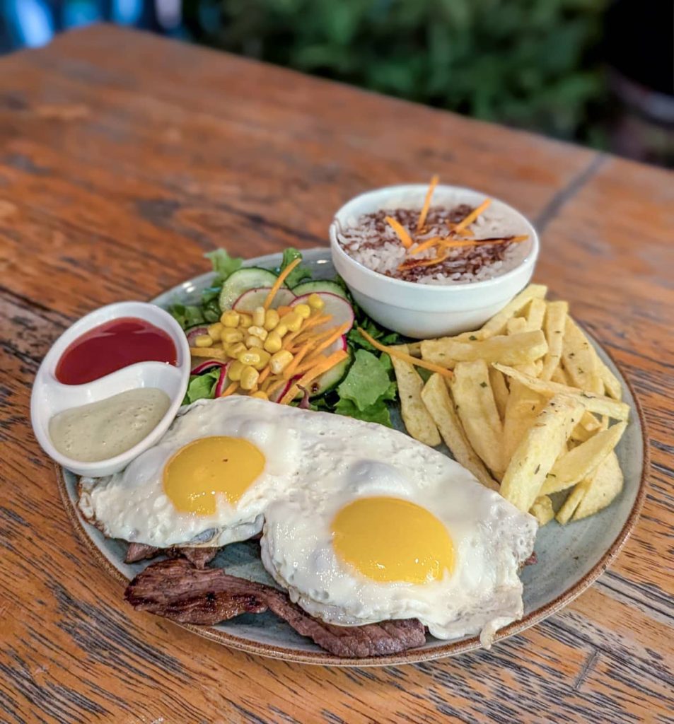 A plate of Ecuadorian churrasco with grilled beef topped with two fried eggs, served with fries, a bowl of rice, salad, and dipping sauces. It's a filling and popular Ecuadorian food enjoyed for breakfast and dinner.