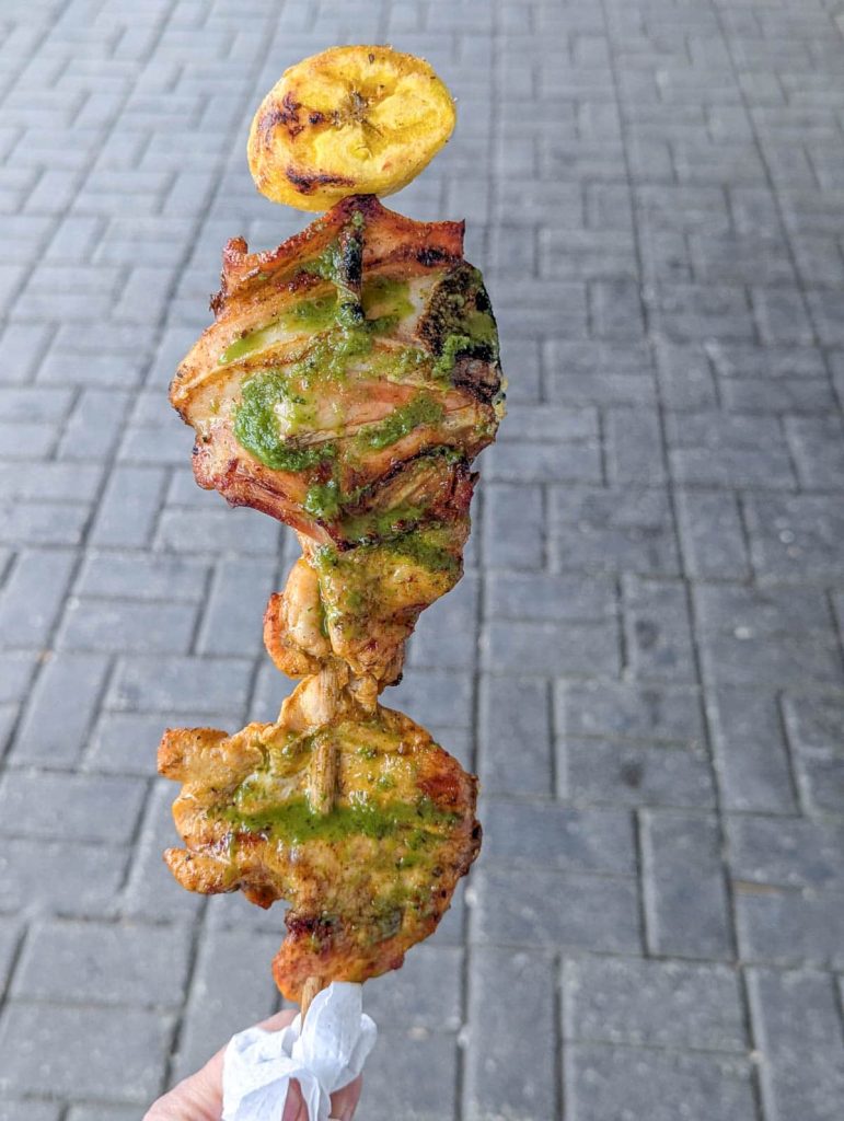 Grilled chicken skewer topped with a slice of grilled plantain, seasoned with a green herb sauce, held against a paved street background in Ecuador. Chuzo is a popular Ecuadorian street food.