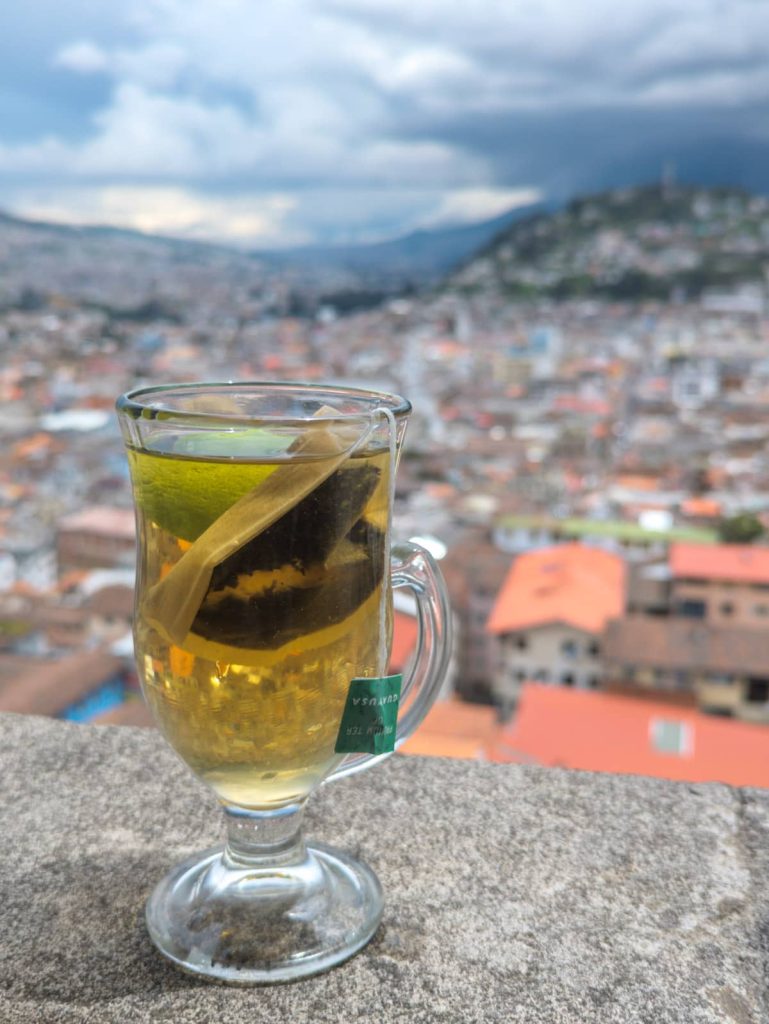 A clear glass mug of guayusa tea, a Amazonian drink of Ecuador. The mug contains a tea bag steeping in golden liquid, perched on a ledge overlooking a cityscape in Quito Ecuador.