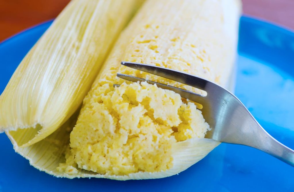 A fork cuts through a soft, freshly steamed humita, an Ecuadorian snack wrapped in corn husk sitting on a bright blue plate.