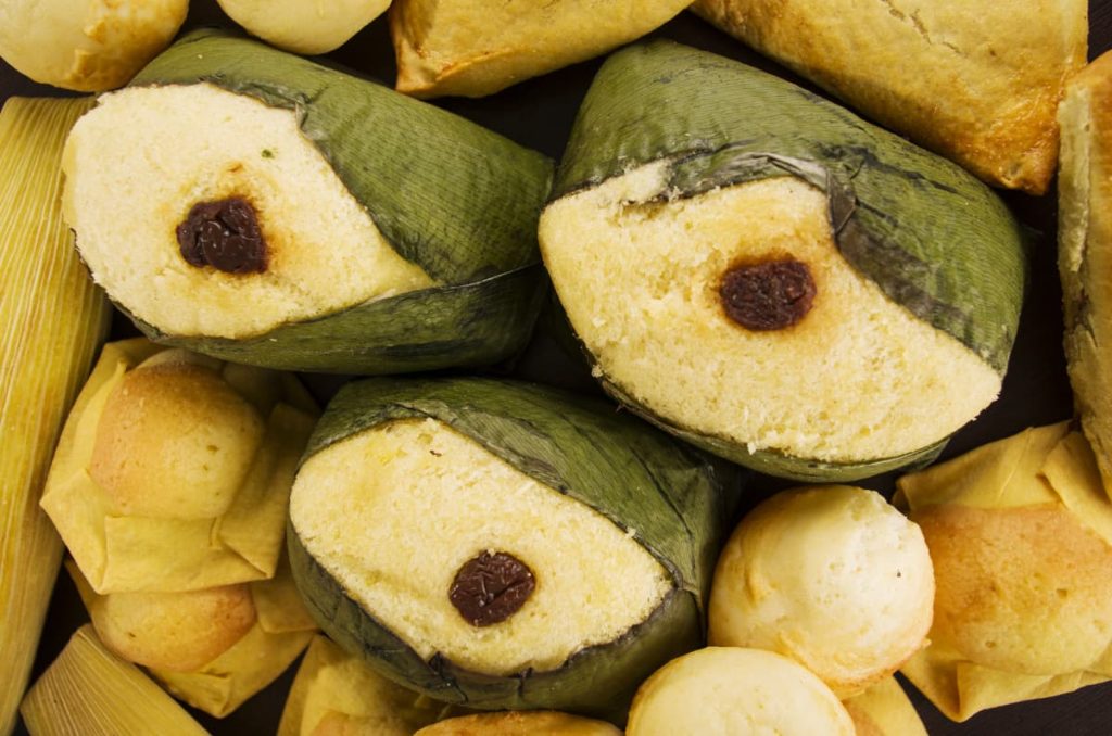 Quimbolitos, a street food in Ecuador that is wrapped in achira leaves and each topped with a raisin are surrounded by other traditional snacks.
