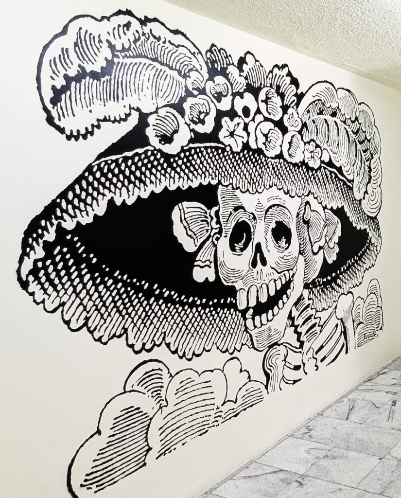 Large black-and-white mural reproducing the original La Catrina by José Guadalupe Posada — a grinning skeleton in a wide feathered hat — painted on a white interior wall of a museum in Mexico City.