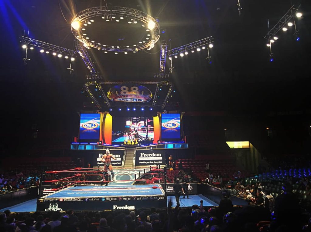 A luchador stands on the middle rope while igniting the crowd before lucha libre in Mexico City. Behind are large television screens to show the wrestling match. Above are track lighting including a large ring of light to light up the stage.