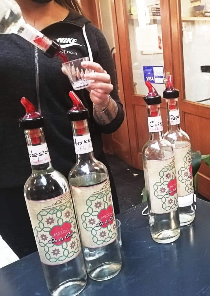 A woman pours mezcal into a glass during a lucha libre tour in Mexico City. On the table in front of her are four other bottles of mezcal.