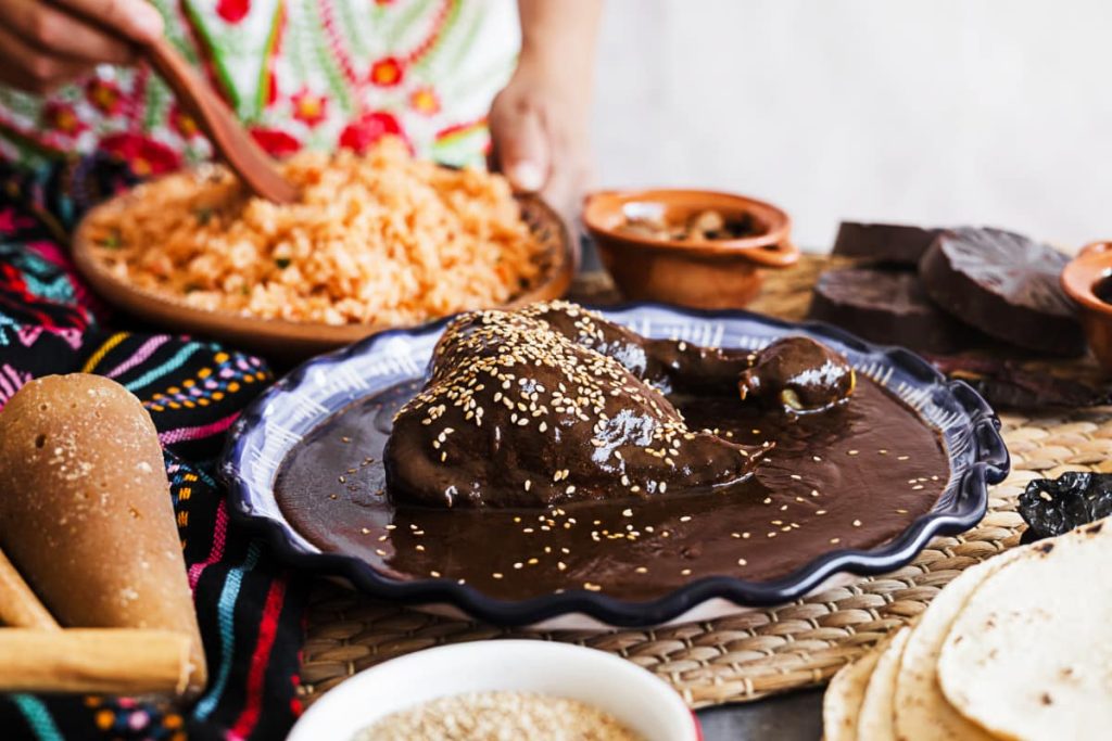 A plate of mole negro topped with sesame seeds sits on a plate during a private cooking class in Mexico City. Surrounding the plate are ingredients to make mole and in the background, hands use a wooden utensil to stir a plate of rice.