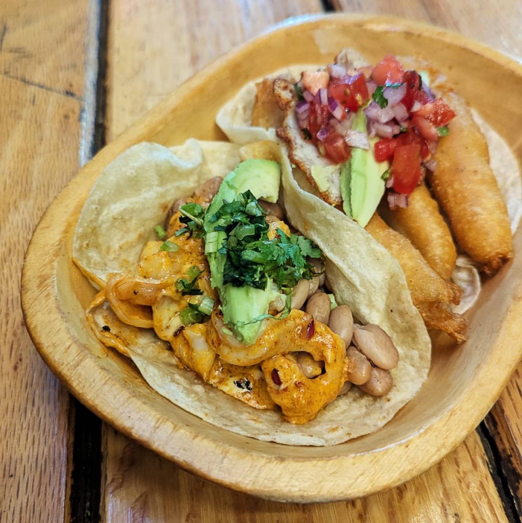 A creamy shrimp taco topped with avocado, cilantro, and beans sits next to a Baja-style fried fish taco topped with pico de gallo.