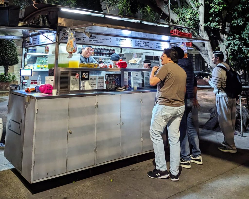 Customers order from Los Caramelos taco cart in Mexico City lit up at night, with a simple stainless steel car and a simple menu in black lettering with a white background.