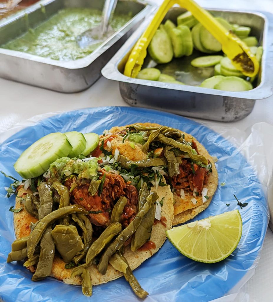 Mixiote lamb tacos topped with grilled nopales, onion, and green salsa, with a lime wedge on the side. Trays of green salsa and cucumber slices sit in the background.