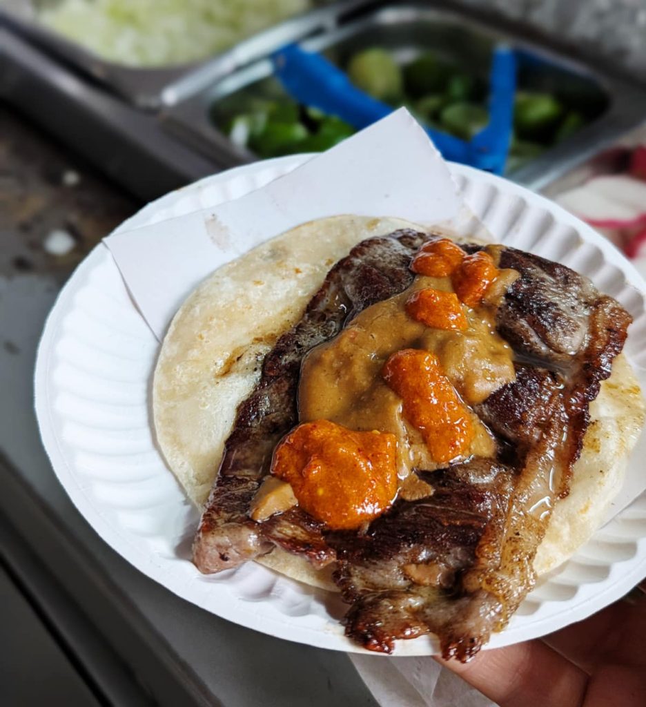 A grilled asada taco in Mexico City on a flour tortilla, topped with two type of salsa, served on a paper plate. In the background are trays of limes, sliced radishes, and onions.