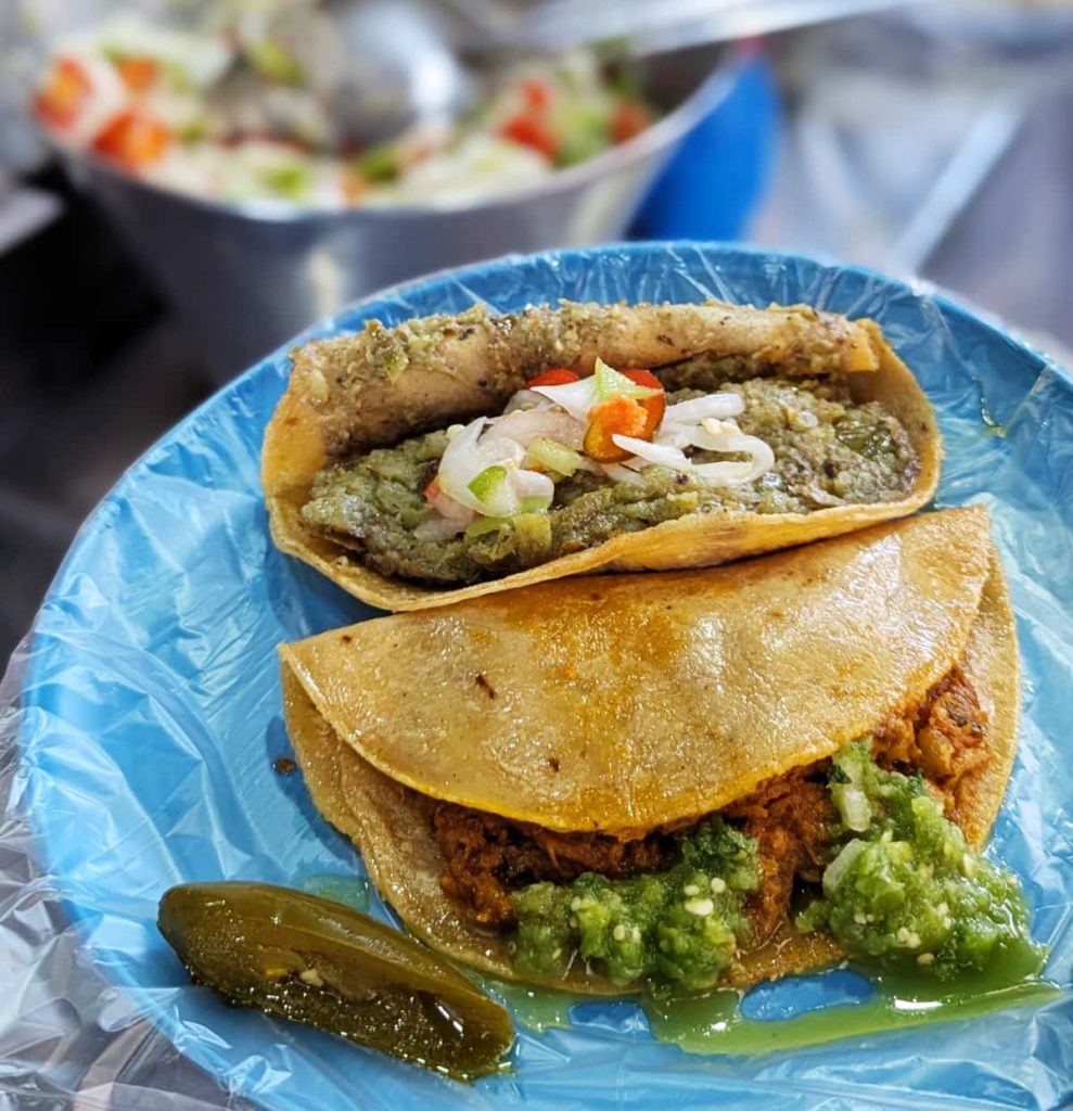 A plastic plate with two tacos de canasta in Centro Mexico City. One is topped with a vibrant green salsa. A pickled jalapeño sits on the blue plate alongside the tacos.