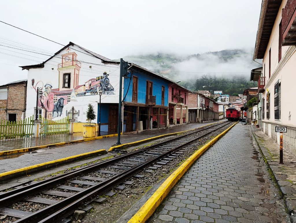 In Alausí, train track run through one of the streets next to a row of pastel colored buildings, one with a train mural that also features a bull. In the background a red train car sits on the tracks and misty clouds encapsulate the mountains.