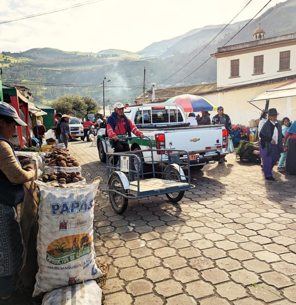A vendor pedals a cargo tricycle through the Alausí Sunday market, past sacks of potatoes with mountains rising behind.