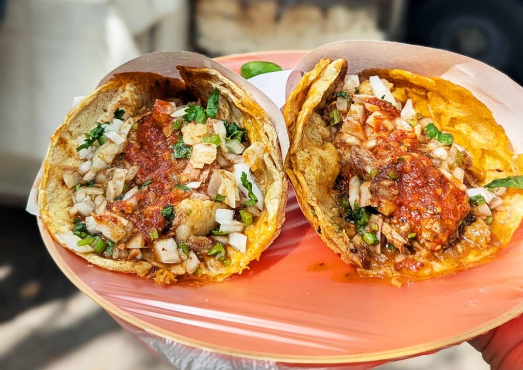 Two birria tacos slightly rolled and wrapped in butchers paper on a plate covered in plastic. The cripsy double tortillas are filled with stewed beef birria and topped with onions, cilantro, and a red salsa.