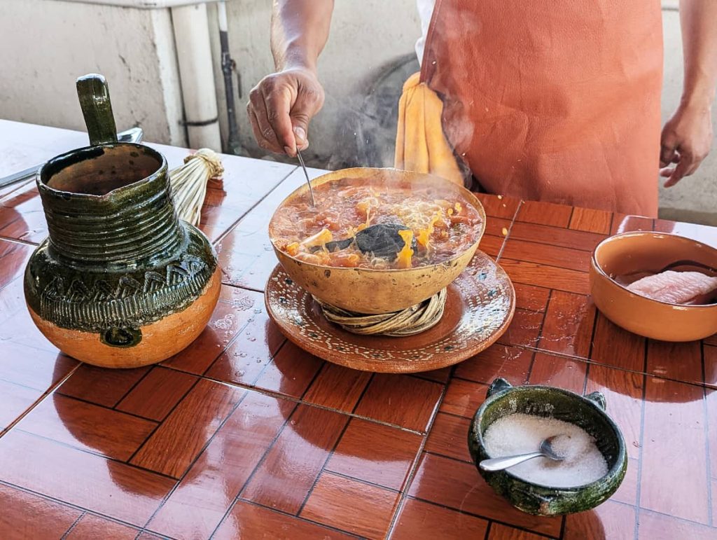 Broth boiling in a gourd bowl with a hot rock inside, as a cook in an orange apron stirs the Caldo de Piedra, a unique Mexican dish. On the counter are a green clay pitcher, bowl of salt, and a piece of raw fish in a bowl.