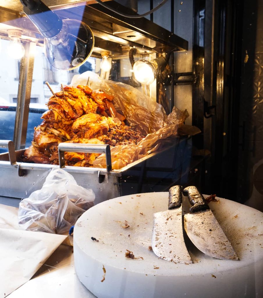 A pile of carnitas in a metal display tray under heat lamps, with two large butcher knives resting on a circular white plastic cutting board, a classic type of pork taco in Michoacán, Mexico.