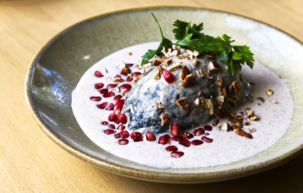 Chile en nogada, one of the most beloved and unique Mexican dishes features a stuffed poblano covered in creamy white walnut sauce, pomegranate seeds, chopped nuts, and parsley on a gray ceramic plate.