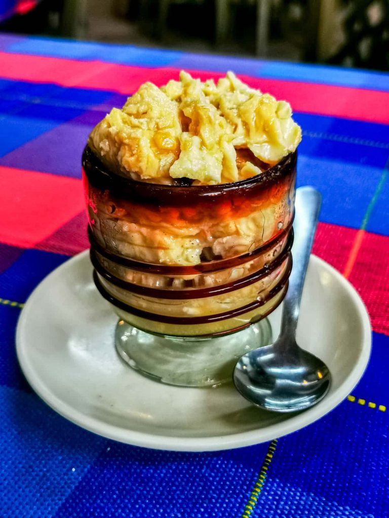 One of the most unique desserts in Mexico, hongos zamoranos is heaped in an amber glass cup with brown rings on a white saucer with a spoon, over a pink and blue tablecloth. This curdled milk dessert resembles scrambled eggs.