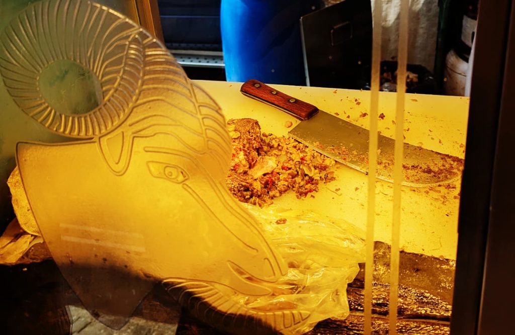 A glass case etched with the head of a ram with curled horns frames a cutting board where chopped barbacoa de borrego sits next to a cleaver with a wooden handle, lit in warm amber light.
