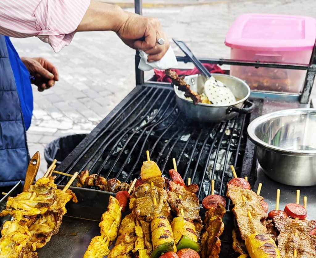 Mixed meat skewers of chicken, pork, sausage, and pineapple piled next to a grill at a street cart in Alasuí Ecuador as a man's hand places a skewer on the grill.