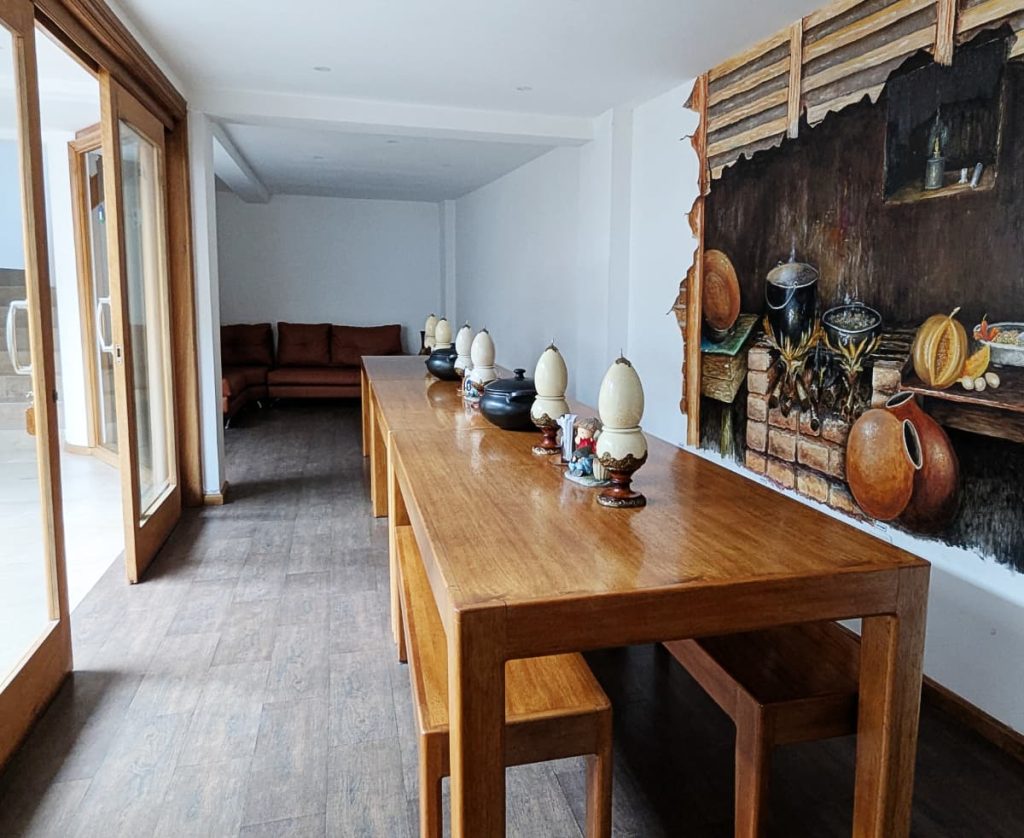 A long wooden table and benches In the dining room at Community Hostel Alausí. A rustic kitchen mural is painted on the wall beside it and glass doors open to a courtyard on the other side.