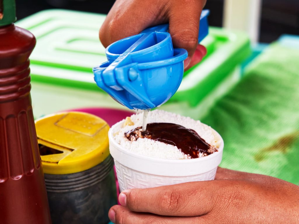A styrofoam cup packed with corn esquites is topped with a thick layer of crumbled white cheese and dark salsa while a hand squeezes lime juice on top using a blue plastic citrus juicer.