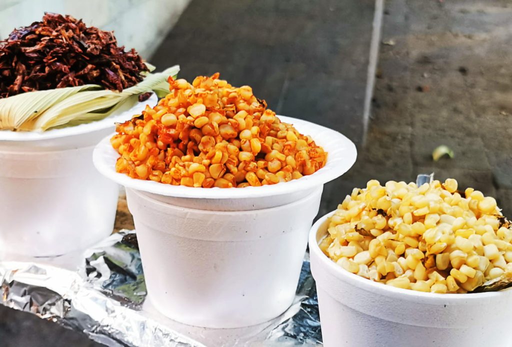 Three styrofoam cups of lined up at a street cart: one piled with corn esquites with red chili, one plain esquites, and the other with toasted grasshoppers, called chapulines.