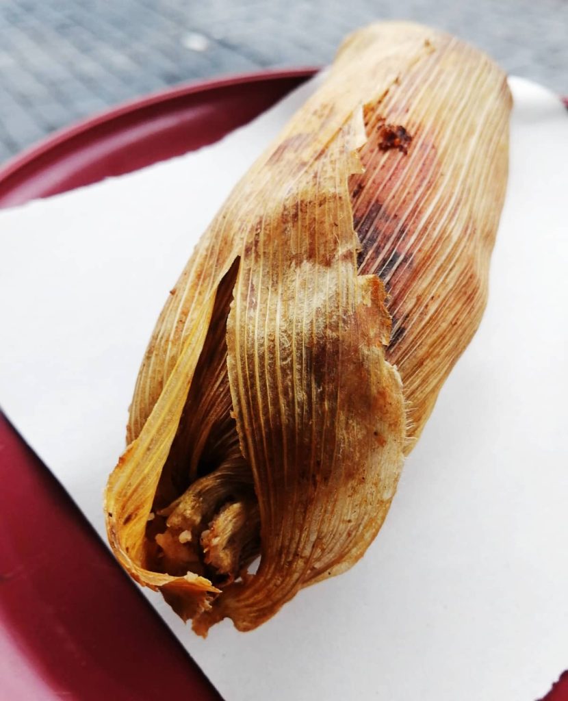 A single corn husk tamale stained with red sauce and slightly open at one end, resting on white paper on a red plastic plate. A tamale in its classic form.