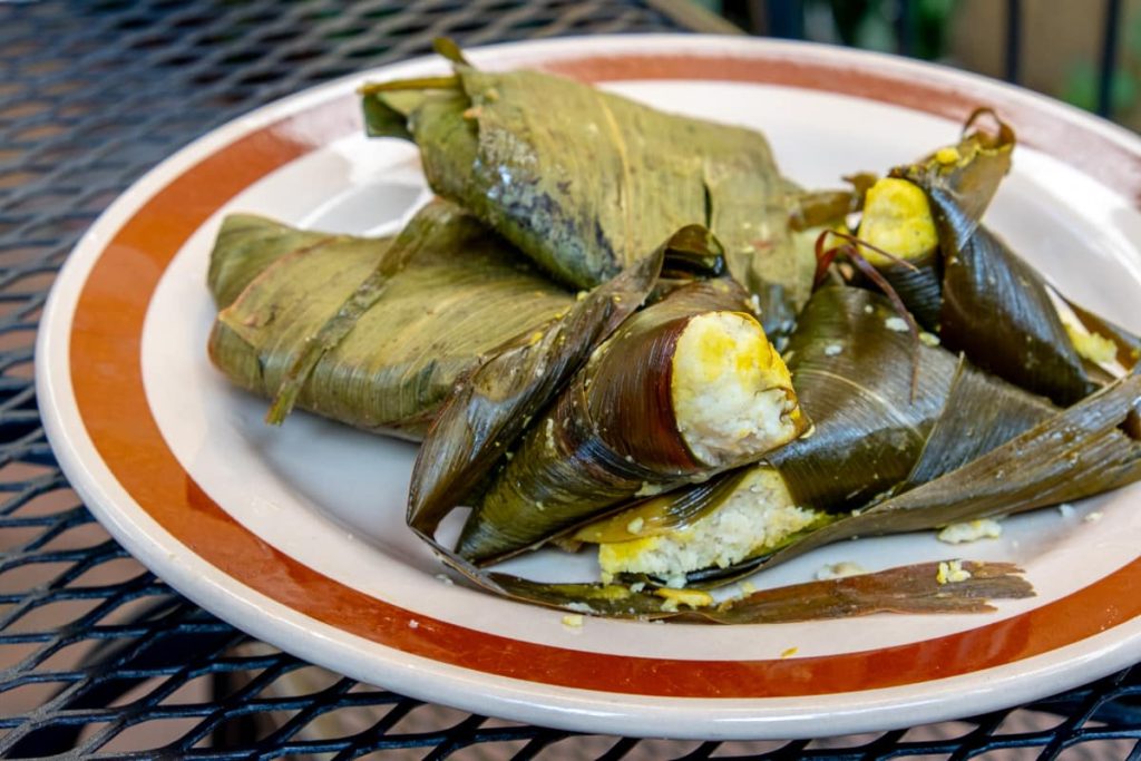 A plate full of a triangular type of Mexican tamale called corundas from Michoacán, Mexico. Pale yellow corn masa is poking through the corners of the triangular tamales which are wrapped in dark green corn leaves.