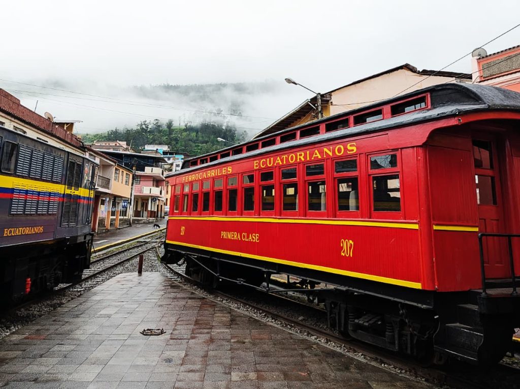 The Devil's Nose Train in Alausí Ecuador parked on a street in the Pueblo Mágico. The train carriage is red with yellow stripes and lettering that says "Ferrocarriles Ecuatorianos, Primera Clase."