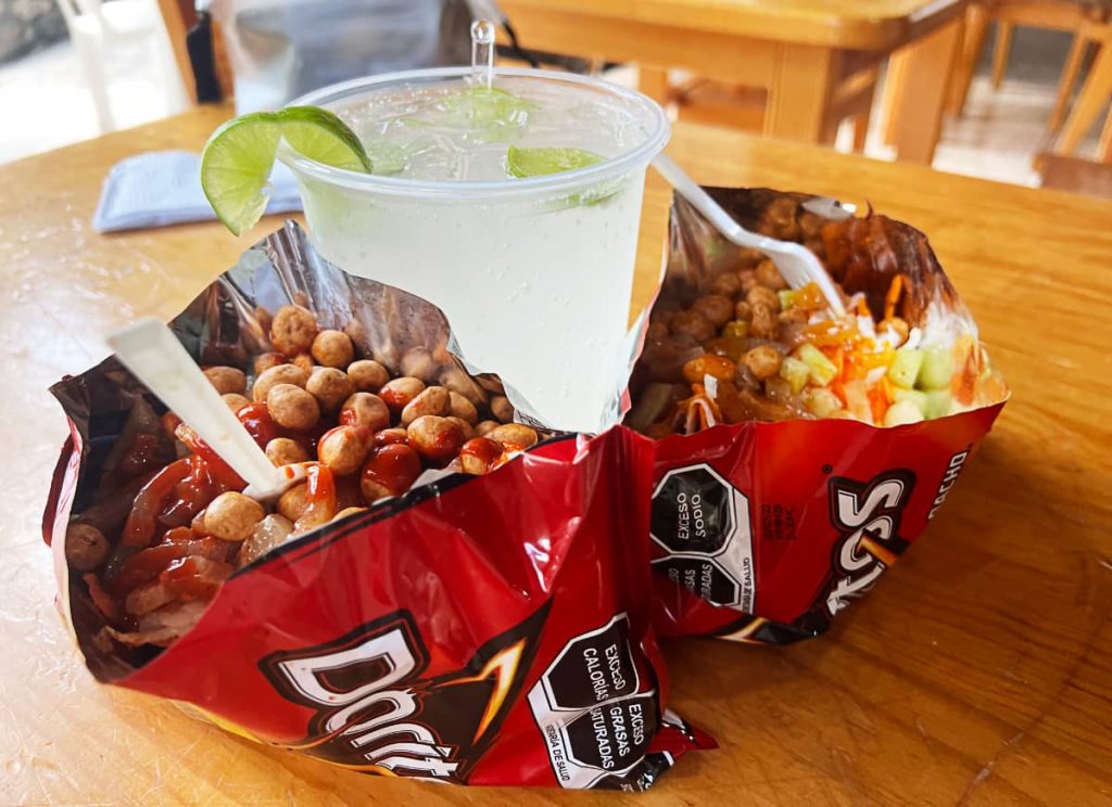 Dorilocos on a wooden table. Two Doritos bags, sliced open and filled with pickled pork skin, Japanese peanuts, cubes of cucumber, and drizzled with a red sauce, sit on a table beside a clear plastic cup of limeade with lime wedges along the rim.
