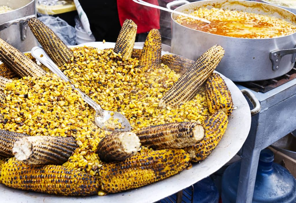 A wide, circular pan with loose grilled corn kernels surrounded by whole charred cobs of corn elotes, with a pot of brothy corn esquites simmering behind it.