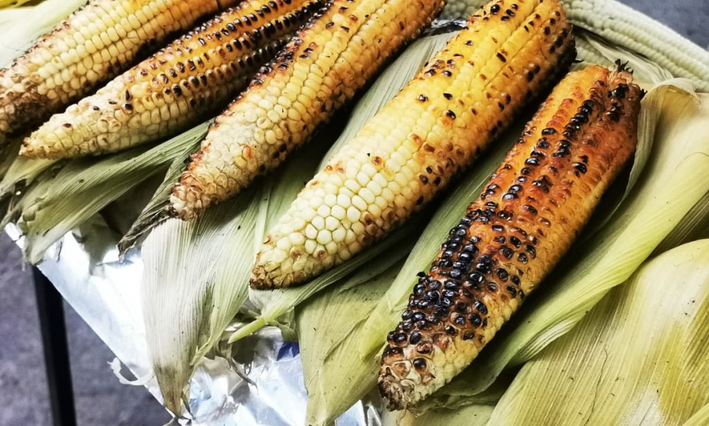 An overhead view of five grilled cobs of corn (elotes), husks peeled back, show kernels charred black and golden brown, resting on a bed of husks with foil underneath.
