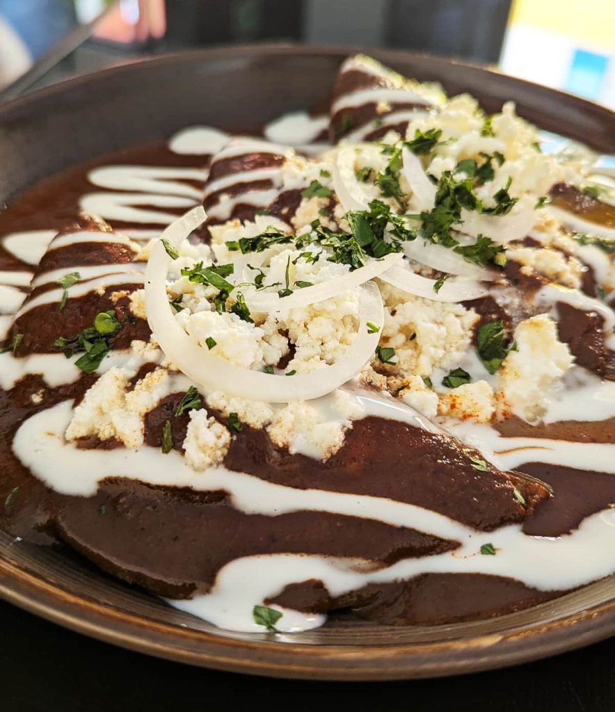 A close up side view of enfrijoladas, a typical breakfast in Mexico. Three tortillas are folded and bathed in a bean sauce. They are topped with cream, fresh cheese, round slices of white onion, and parsley.