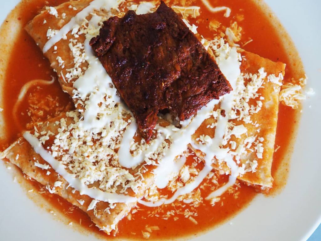 Ann overhead view of a plate of entomatadas, a typical breakfast in Mexico. The white plate has three tortillas folded into triangles and bathed in a red tomato sauce It's topped with cream and shredded cheese. On top is a small piece of meat with red spices.
